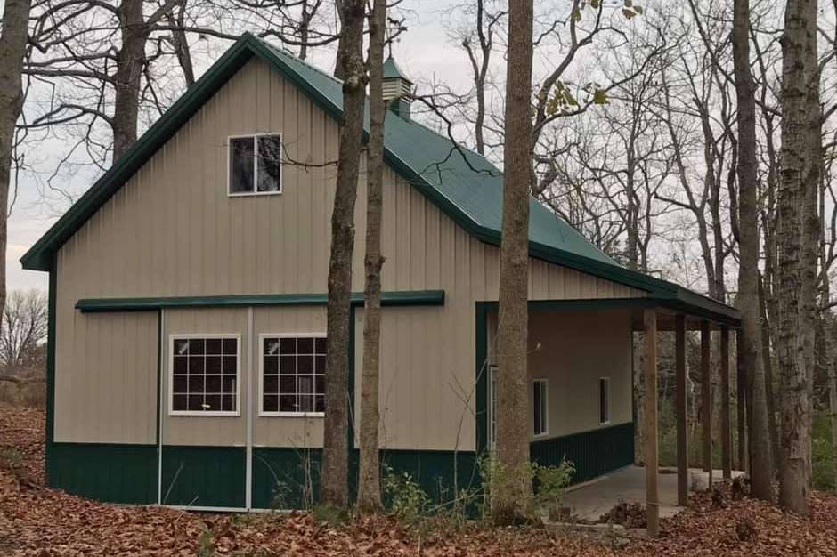 A two-story tan and green pole barn with a metal roof and side porch, nestled among trees in a wooded setting.