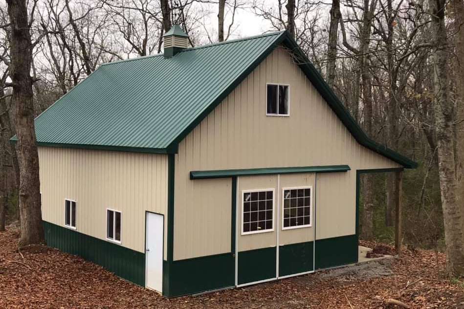 A beige and green metal shed with a matching green roof, set in a wooded area with fallen autumn leaves on the ground.
