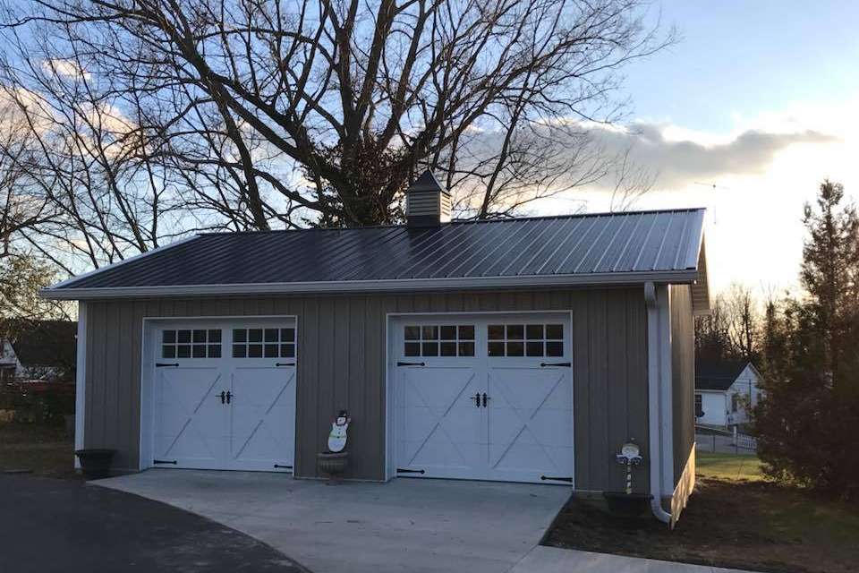 A grey detached two-car garage with white carriage-style doors and a dark metal roof during a sunset.