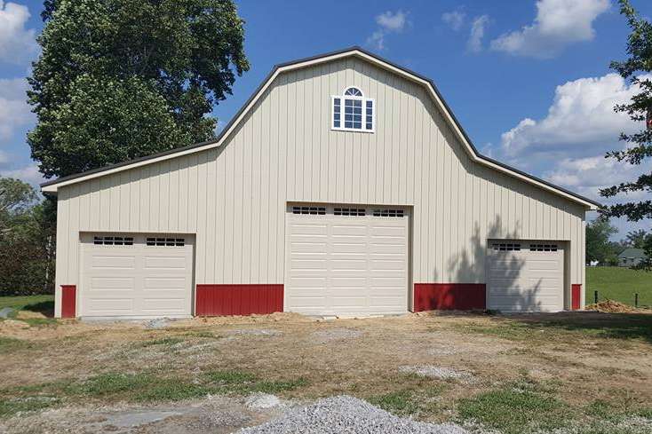 A tan, barn-style building with a red base and three white garage doors, set against a blue sky with green trees.