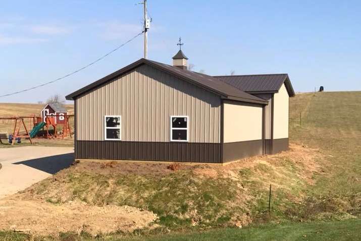 A metal-sided building with beige walls and dark brown trim, set on a small grassy hill near a playground.