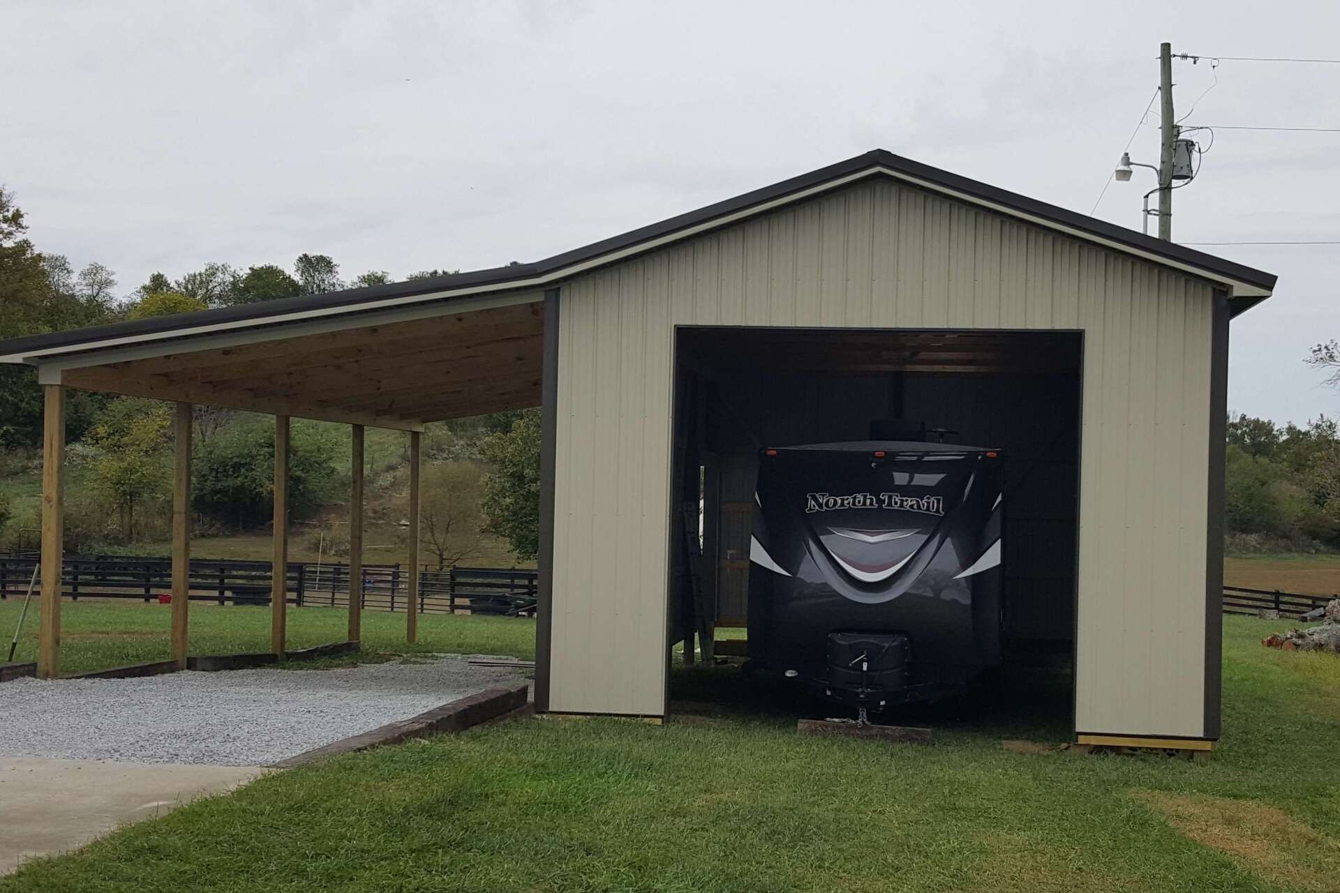 A tan metal garage with an open side carport housing a dark RV on a gravel lot in a rural setting.