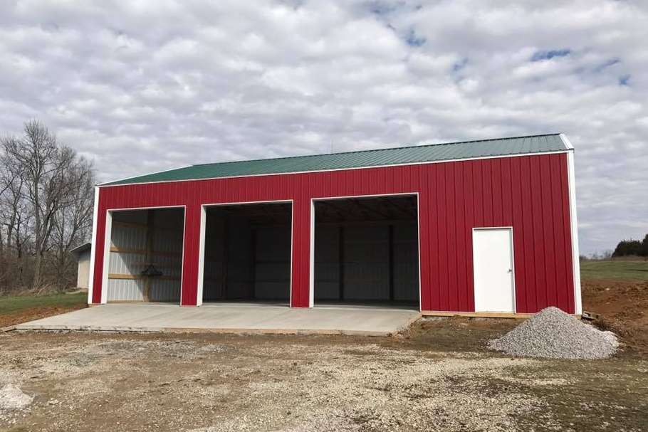 Red metal pole barn with three open garage bays and one white entry door on a gravel lot under a cloudy sky.