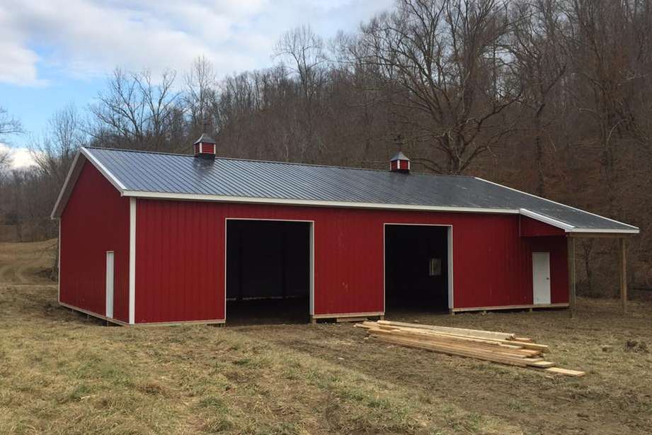 A red pole barn with a dark metal roof, two large open bays, and a side lean-to, set in a rural, grassy field.