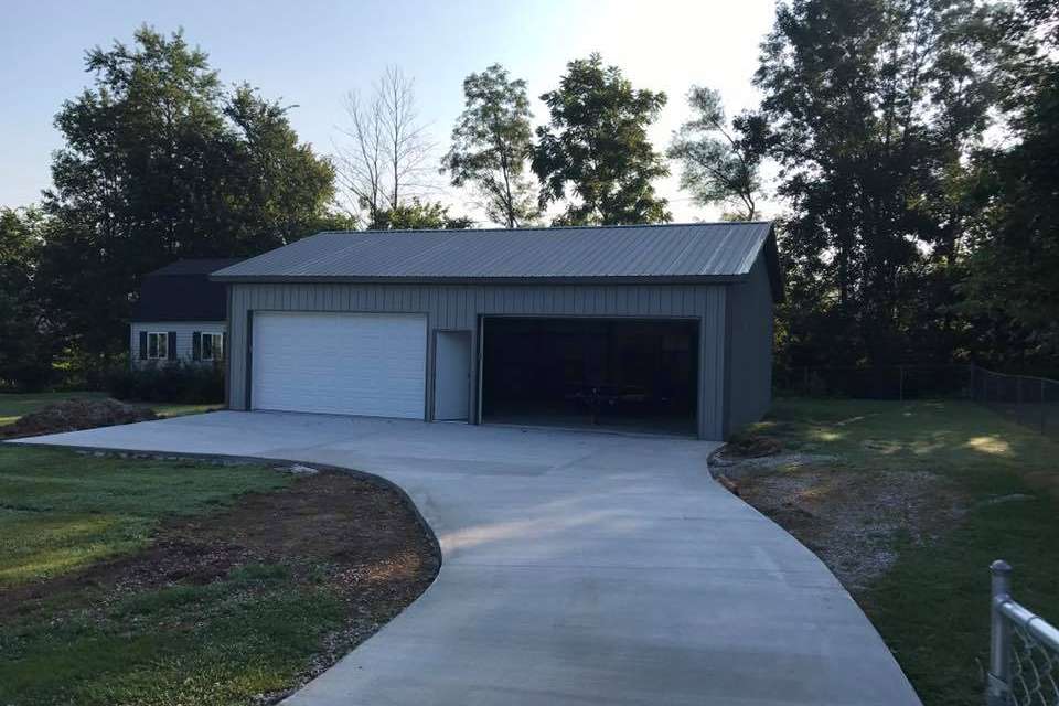 A gray metal garage with one closed white door, one open bay, and a new concrete driveway set among trees.