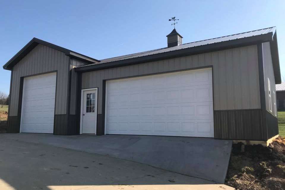 A detached two-tone grey metal garage with a white walk-in door and two white garage doors under a clear blue sky.