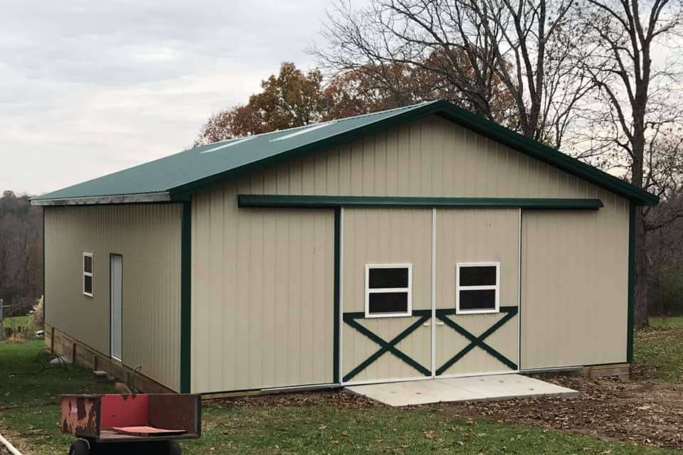 A tan metal garage with a green roof, sliding double doors with windows and X-trim, and a side entry door in a field.