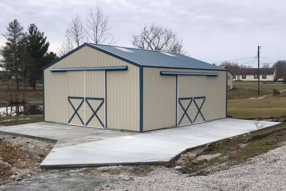 A beige metal barn with blue trim and sliding doors, set on a concrete pad in a rural yard.