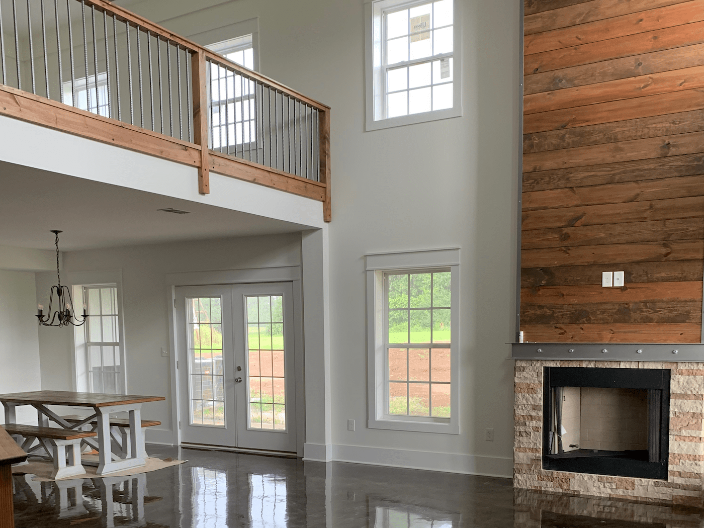 A modern, open-concept living area with a high ceiling, a stone fireplace, wood wall paneling, and a mezzanine balcony.
