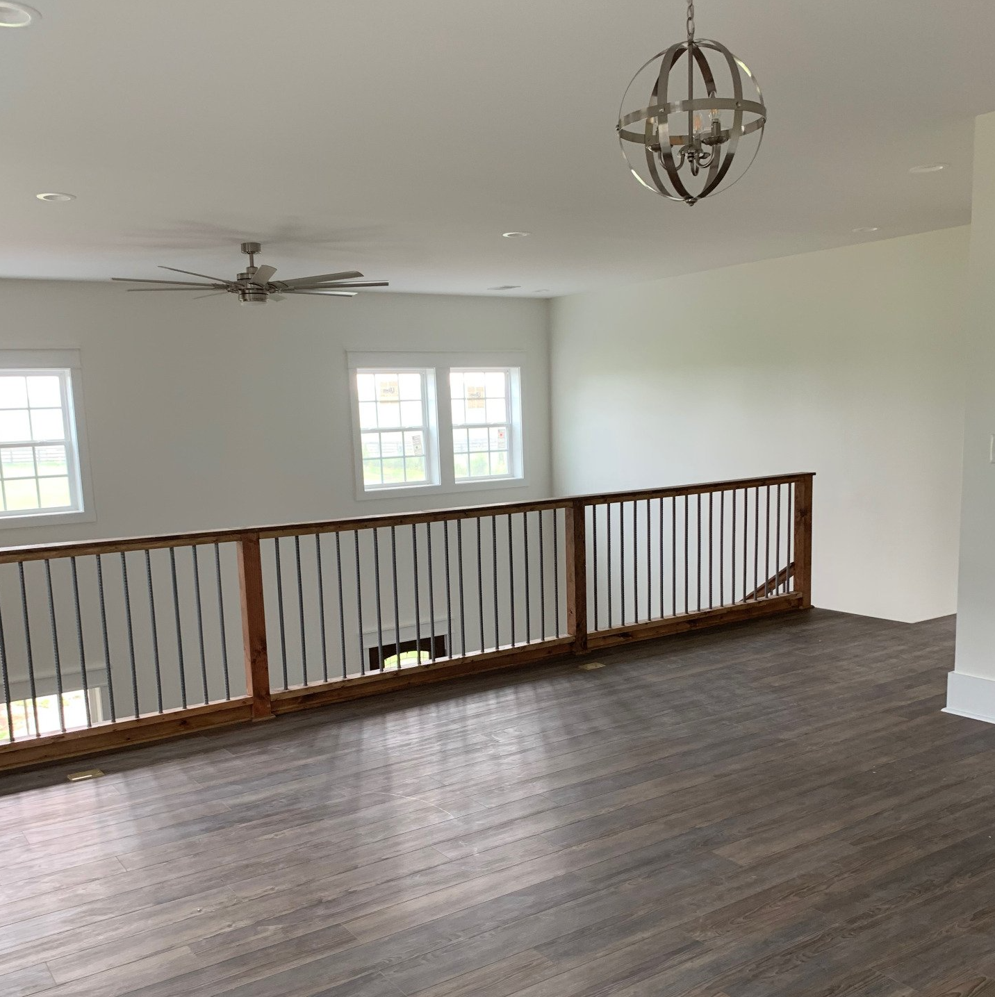 A second-floor loft with gray wood flooring, a wooden staircase railing, two windows, a ceiling fan, and a globe light.
