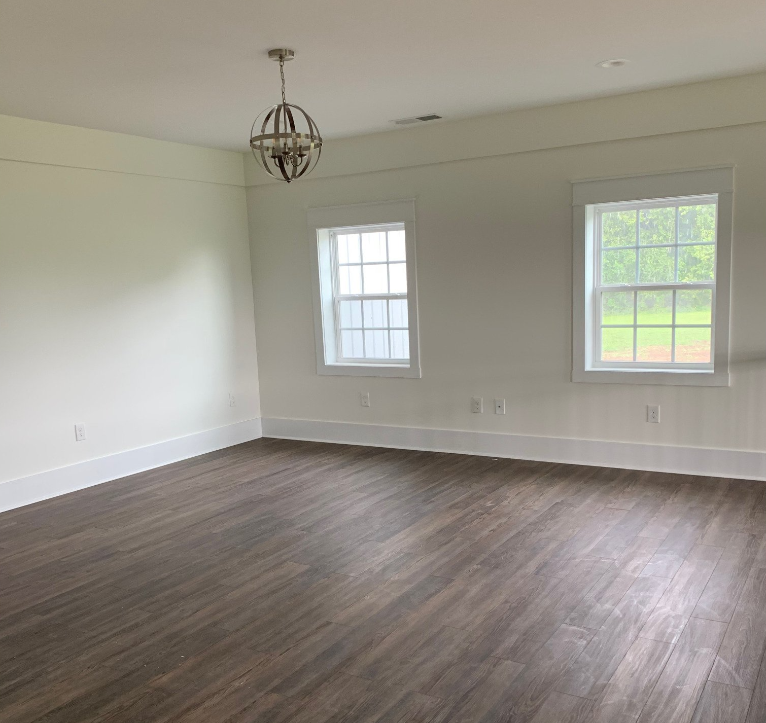 An empty room with light-colored walls, white trim, dark wood-look flooring, two windows, and a metal orb chandelier.