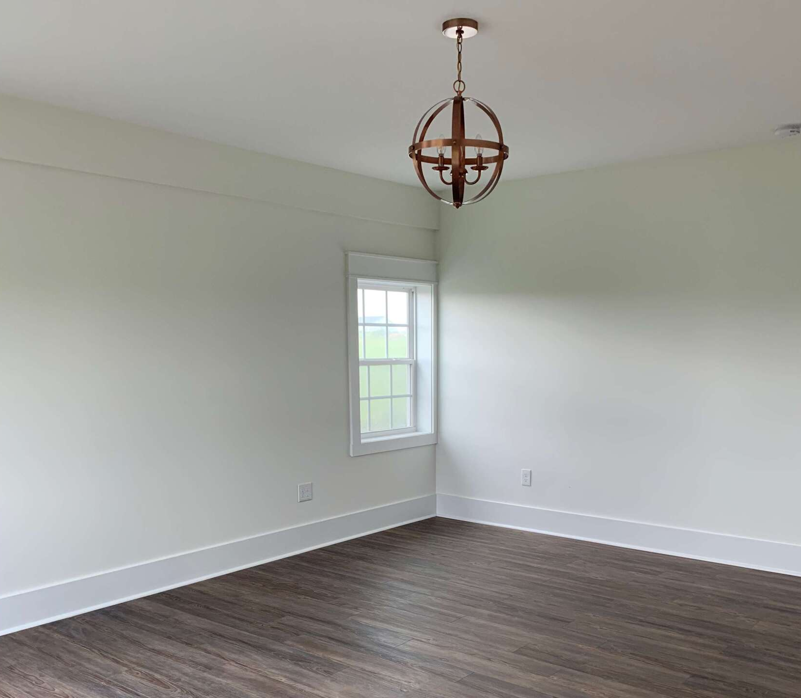 A room with white walls, dark wood-look flooring, a window centered on the left wall, and a rustic orb chandelier above.