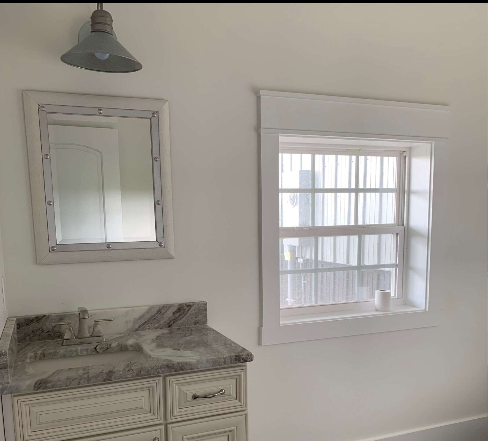 A vanity area with a granite countertop, a framed mirror, a wall-mounted light, and a window with white trim.