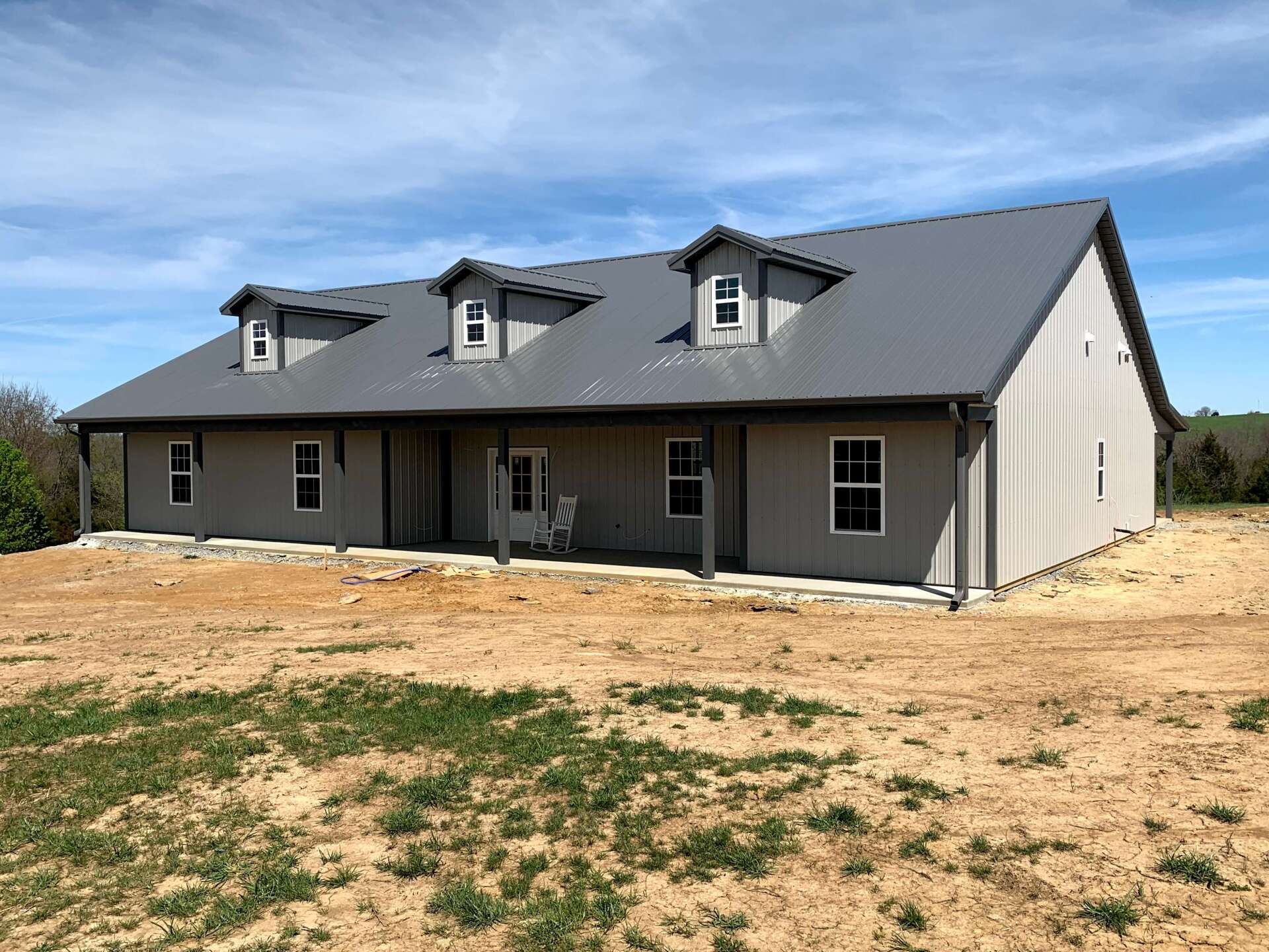 A light gray pole barn house with a dark metal roof, three dormer windows, and a covered front porch on a dirt lot.