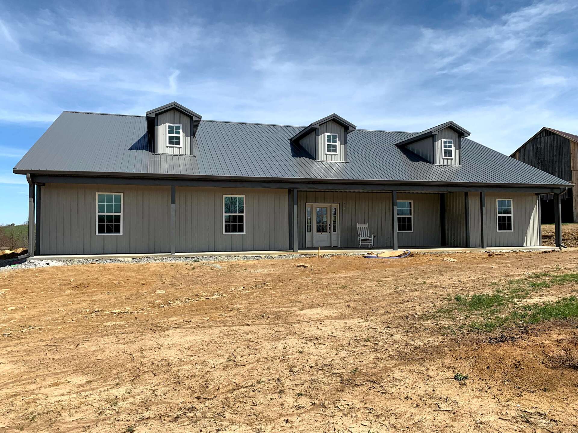 A single-story gray house with a dark metal roof, three dormer windows, and a front porch, set on a dirt lot.
