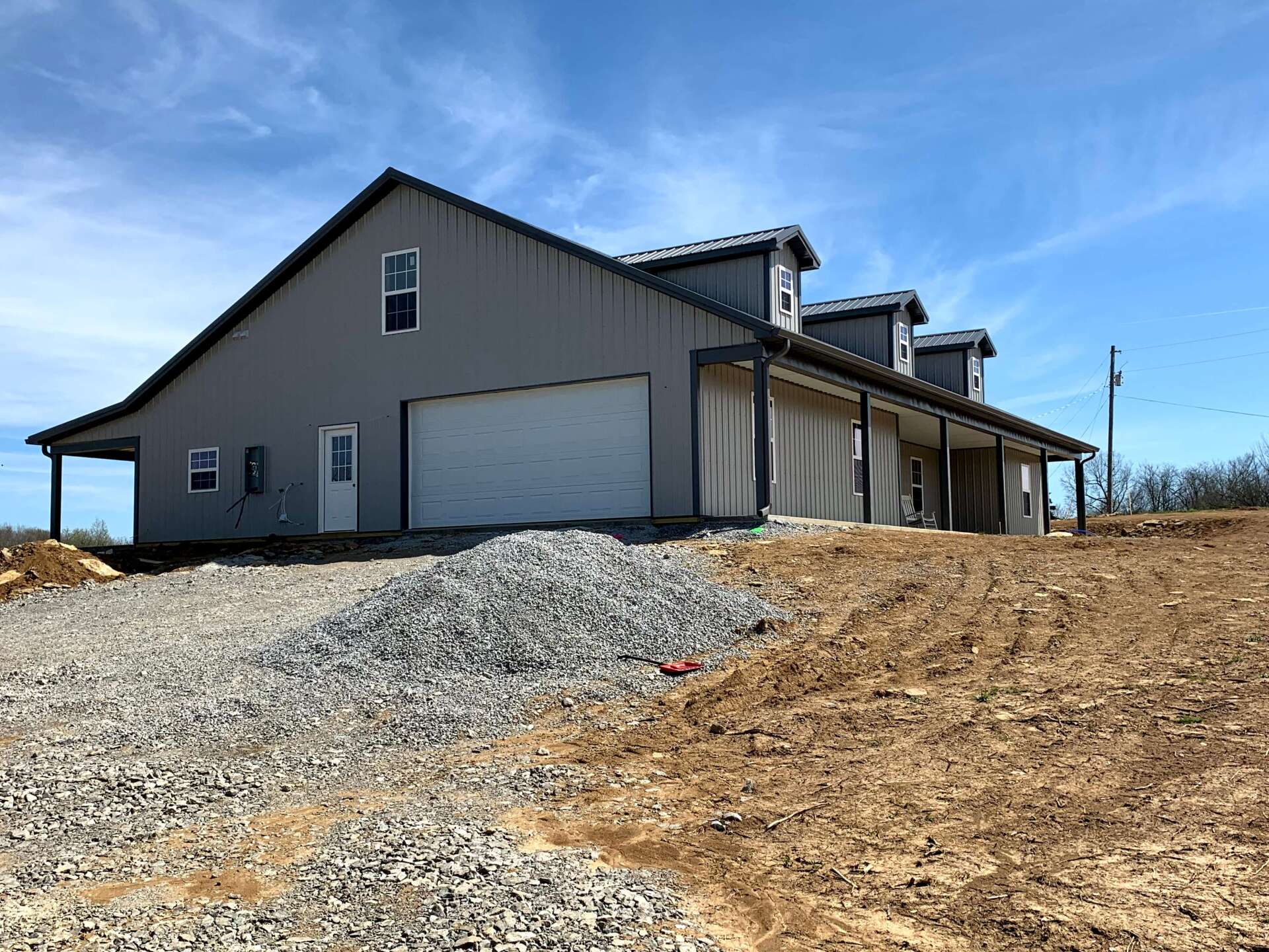 A modern gray metal barndominium with a white garage door, front porch, and dormers sits on a gravel lot under a blue sky.