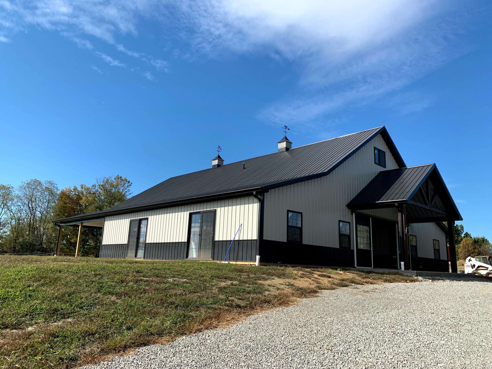 A wide, metal-sided barn with black accents, a dark metal roof, and two cupolas, situated on a gravel lot under blue sky.