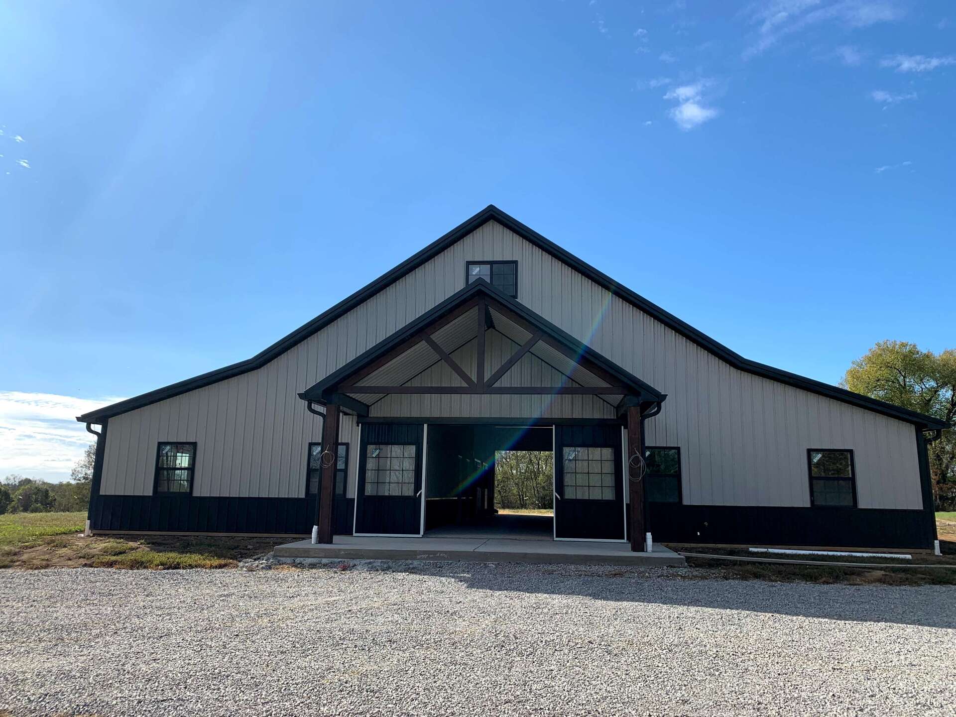 A modern barn with beige metal siding and dark trim features a central covered entryway under a clear blue sky.