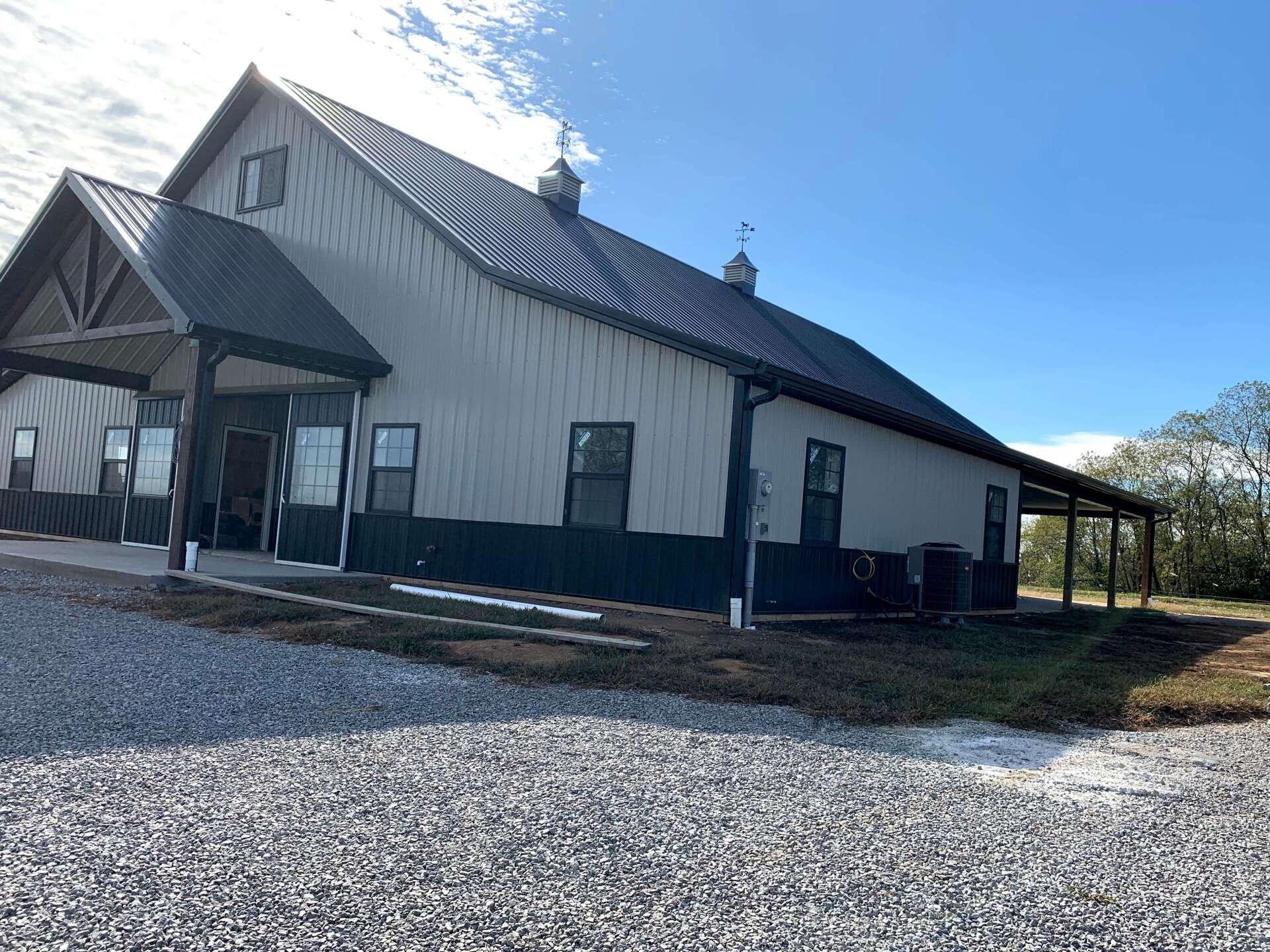 A tan metal-sided building with a dark roof and foundation, featuring a front porch and gravel driveway under a blue sky.