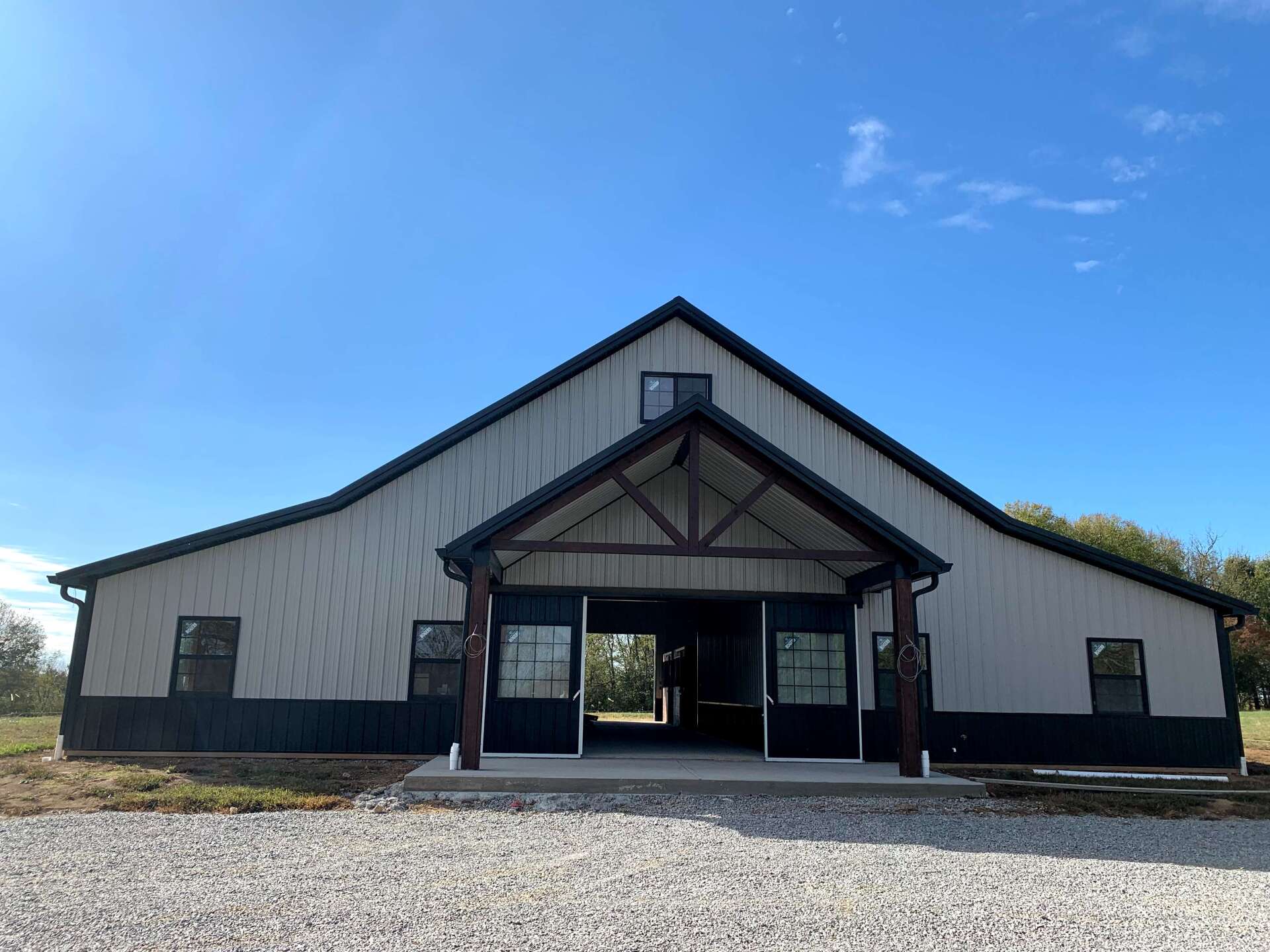 A light-colored metal barn with dark trim, a covered entryway, and a gravel driveway under a clear blue sky.