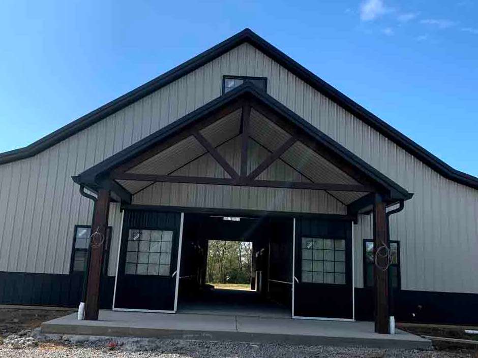 Exterior view of a barn with light metal siding, dark trim, a covered porch, and open doors leading through the structure.