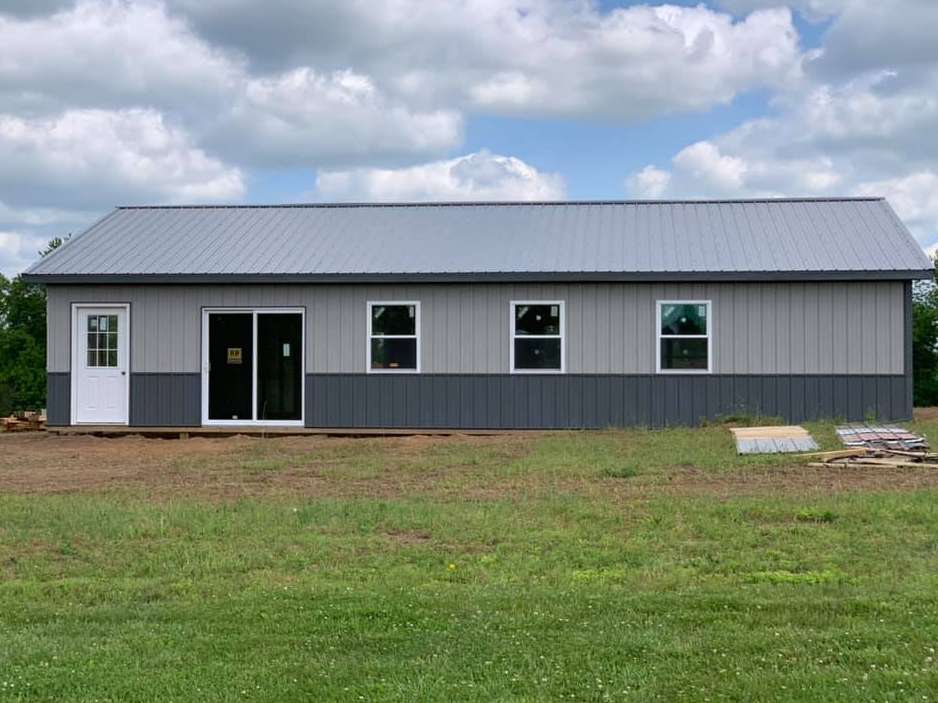 A newly constructed metal pole barn with a gray metal roof, light gray siding, dark gray wainscoting, and white doors.