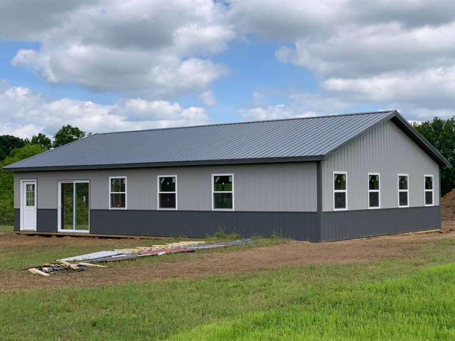 A gray metal building with a dark gray lower trim, a metal roof, several windows, and a white door in a rural field.