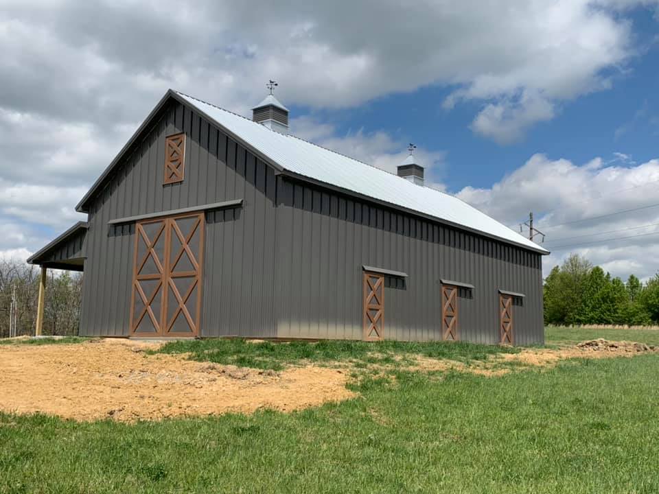 A dark gray barn with a metal roof and wooden sliding doors set in a grassy field under a cloudy blue sky.