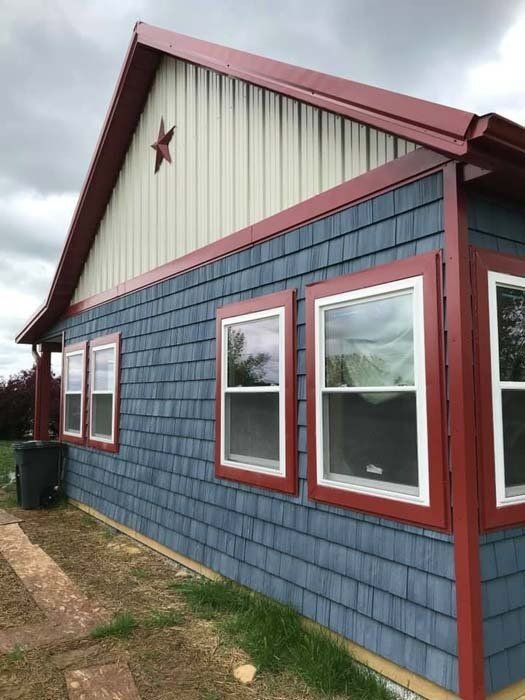 A corner view of a building with blue shingle siding, white window trim, red accents, and beige vertical peak siding.