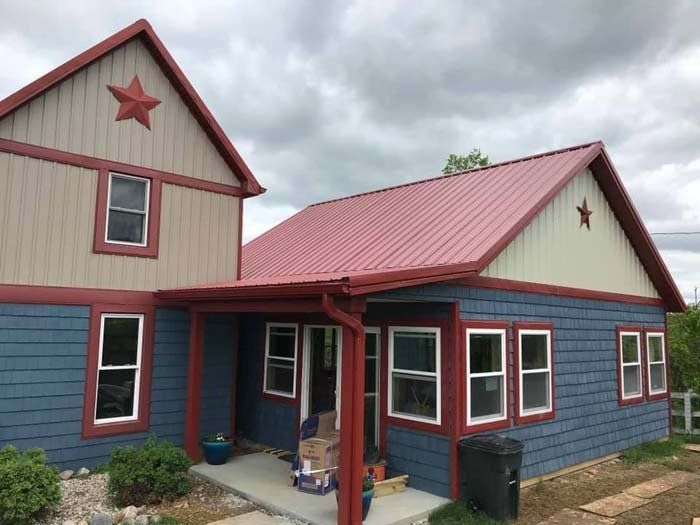 A blue house with a beige second-story loft, a red metal roof, and decorative stars on the exterior gable walls.