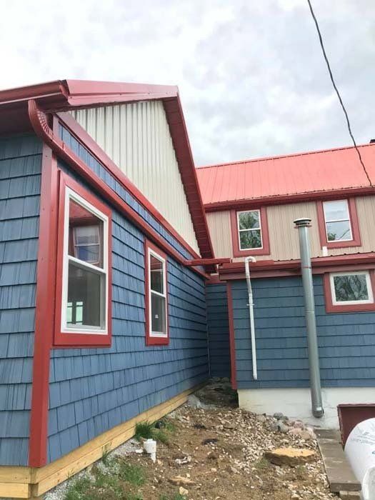 A house exterior featuring blue shingle siding, red trim, a metal roof, and an unfinished gravel foundation area.