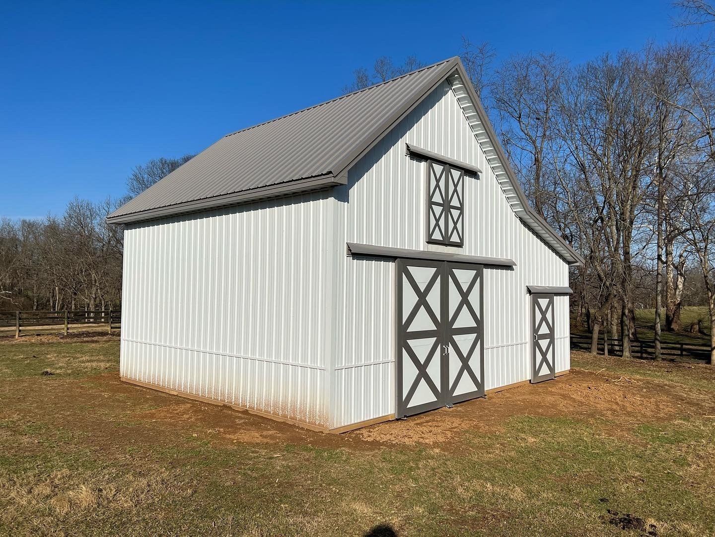 White metal barn with dark grey trim, a sliding double door, a hay door above, and a side entrance in a grassy field.