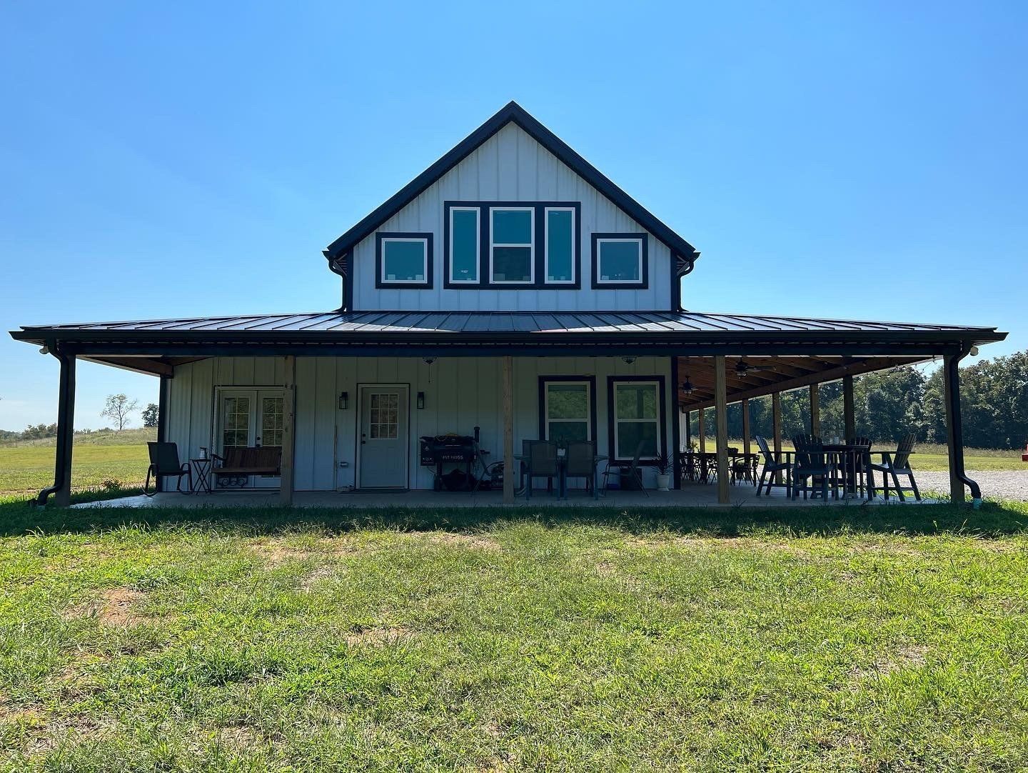 A two-story white farmhouse with a large wraparound porch, dark trim, and a metal roof under a bright, clear blue sky.