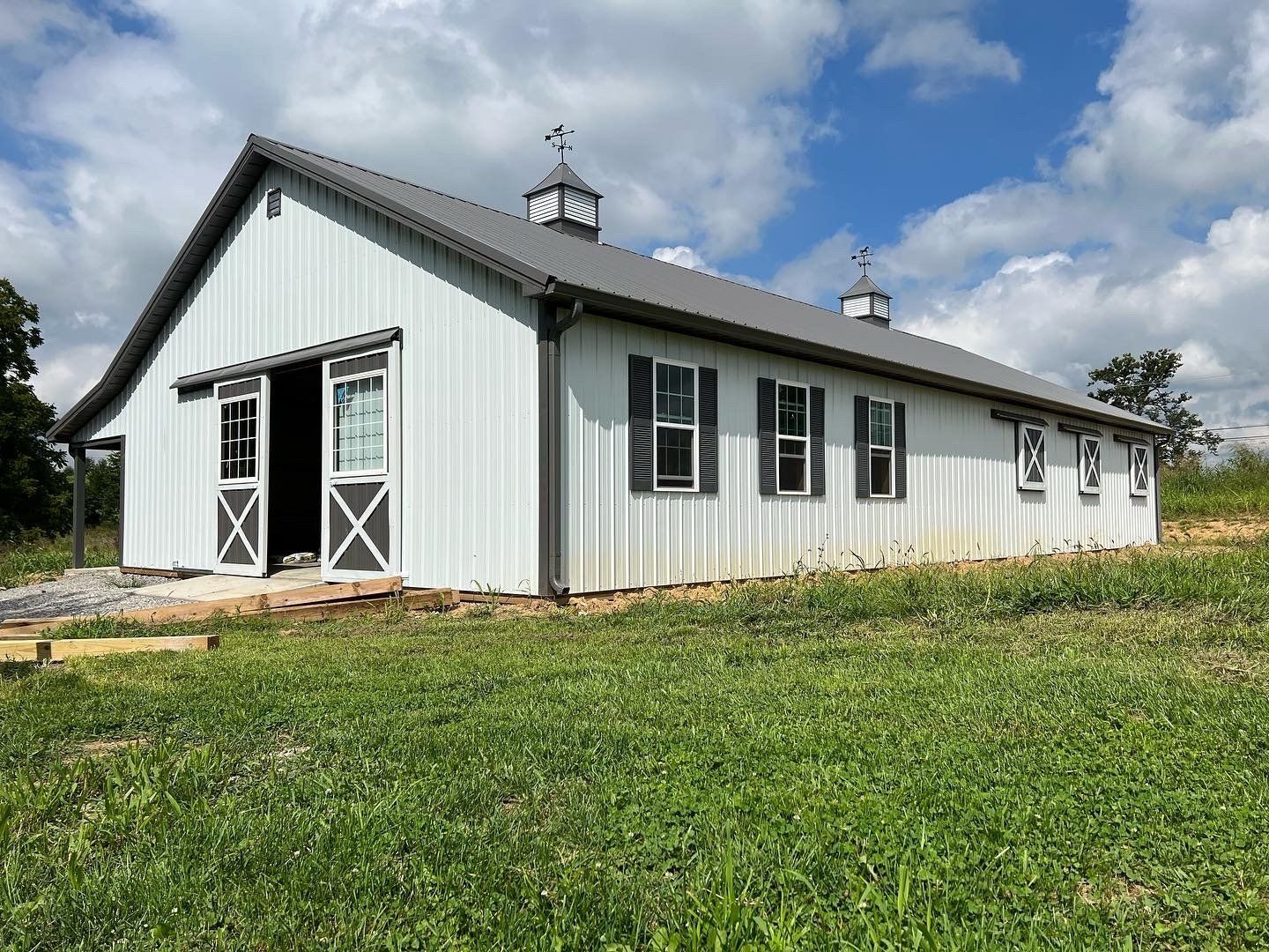A white metal-sided barn with grey trim and a metal roof, featuring dual cupolas, set in a grassy field under a blue sky.