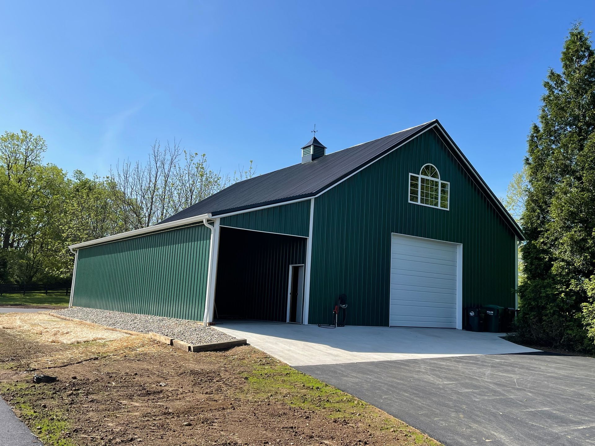 A green metal barn with a black roof, a white garage door, and an open side bay under a clear blue sky.