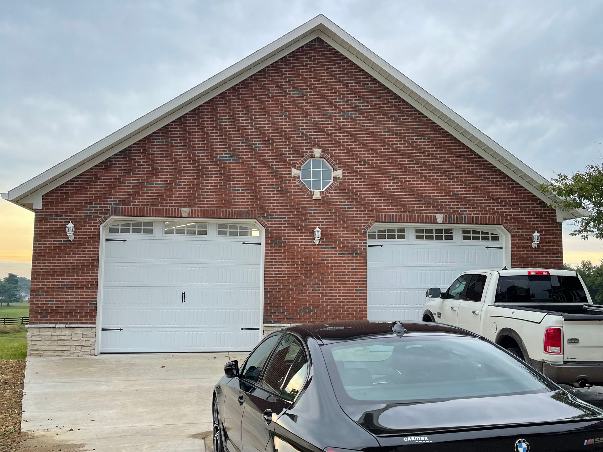 A detached brick garage with two white doors, featuring a decorative central window, with cars parked in the driveway.