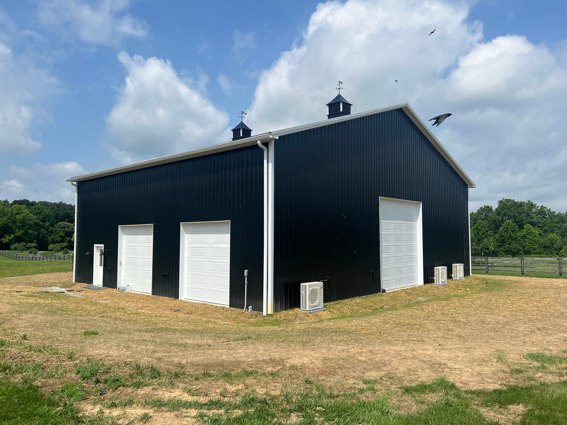 A black metal barn with white garage doors and a man-door, situated in a field under a blue sky with clouds.