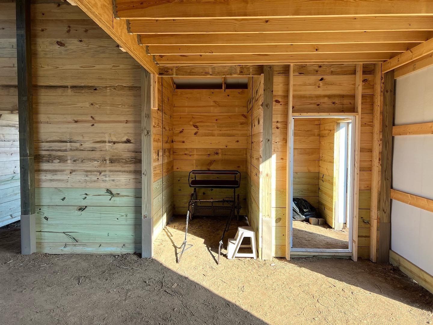An unfinished interior space with light wood paneling, a dirt floor, a small work bench, and an open doorway.