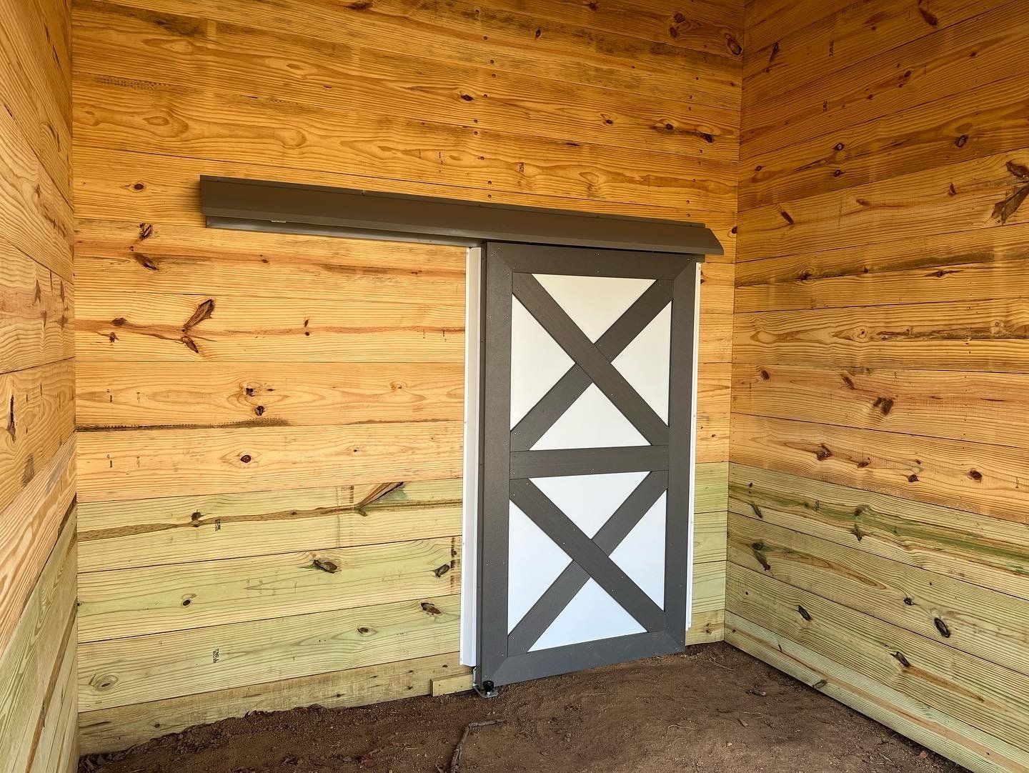 A sliding wooden door with a dark grey X-brace pattern mounted on an interior wall of unfinished wood planks.