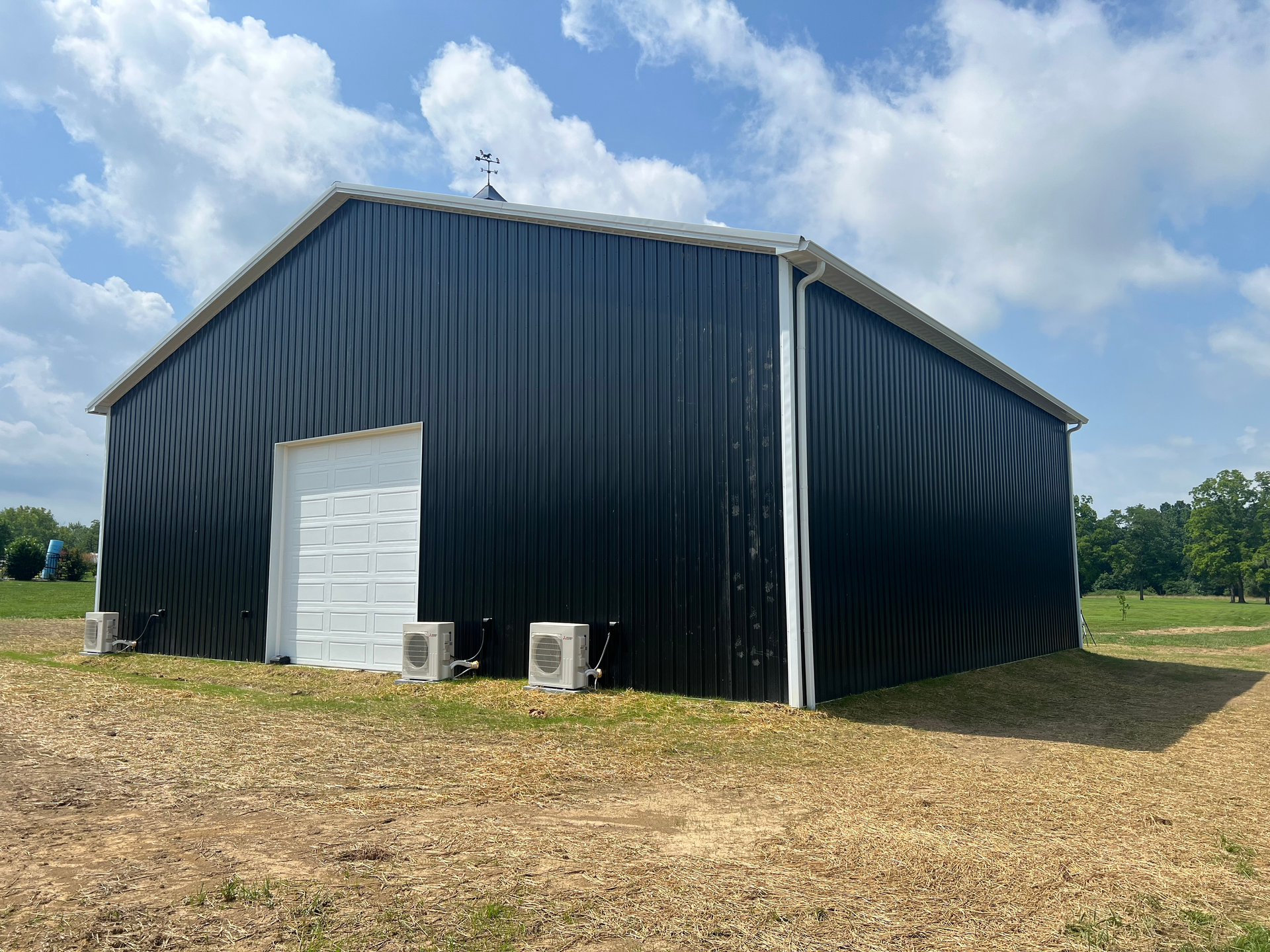A black metal building with a white garage door and two small HVAC units outside in a grassy field under a blue sky.