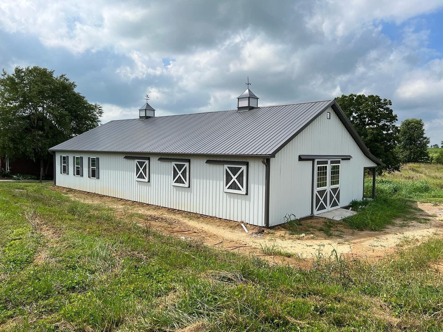 White barn with a dark grey roof and two cupolas, situated in a grassy, partially cleared field under a cloudy sky.