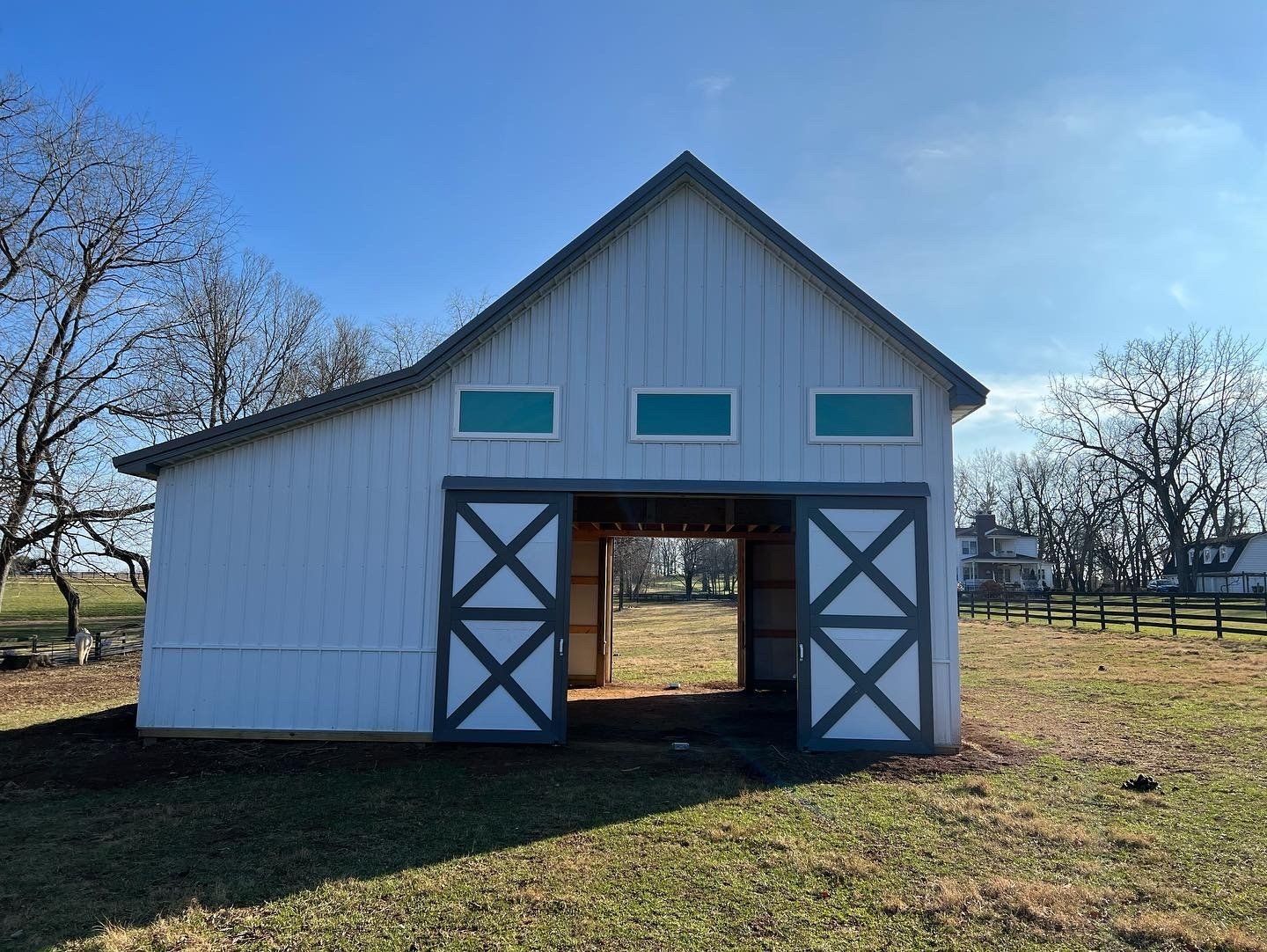 A white barn with a grey roof and X-patterned sliding doors stands in a grassy field under a clear blue sky.