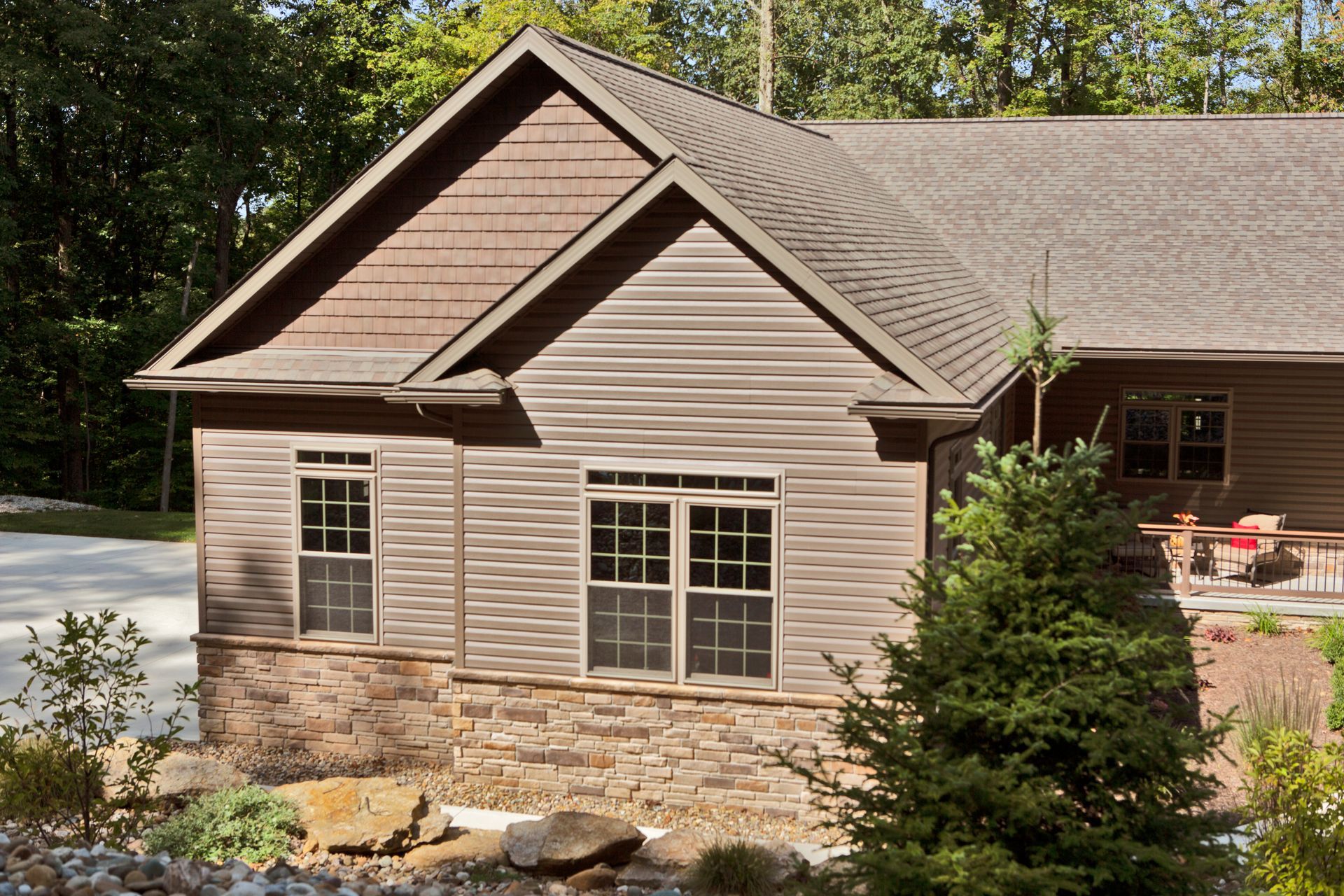 A brown house with wavy horizontal siding, stone veneer base, and a gabled roof set among green trees and bushes.