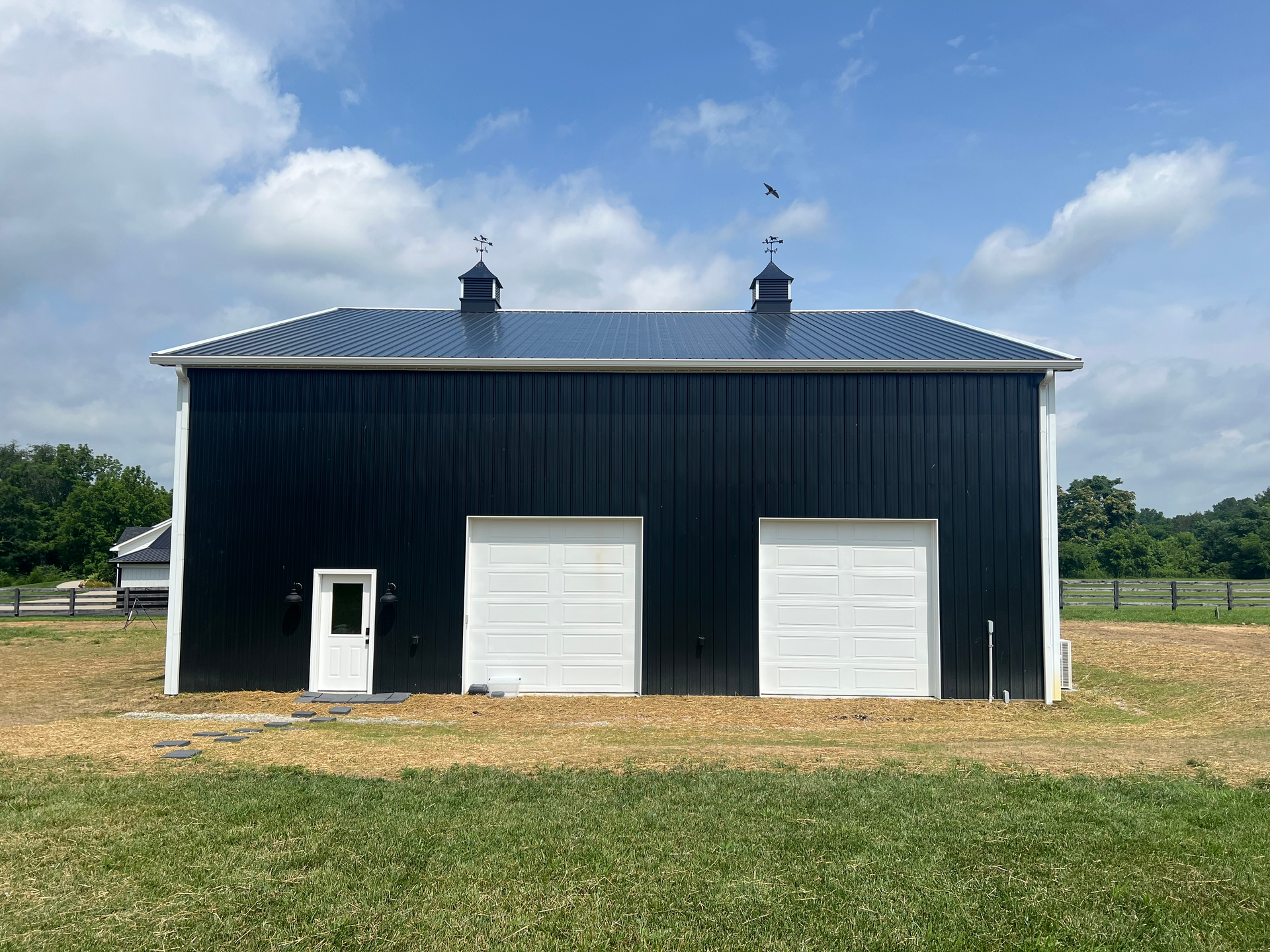A black barn with white doors and a dark metal roof, set against a bright blue sky with clouds over a grassy field.