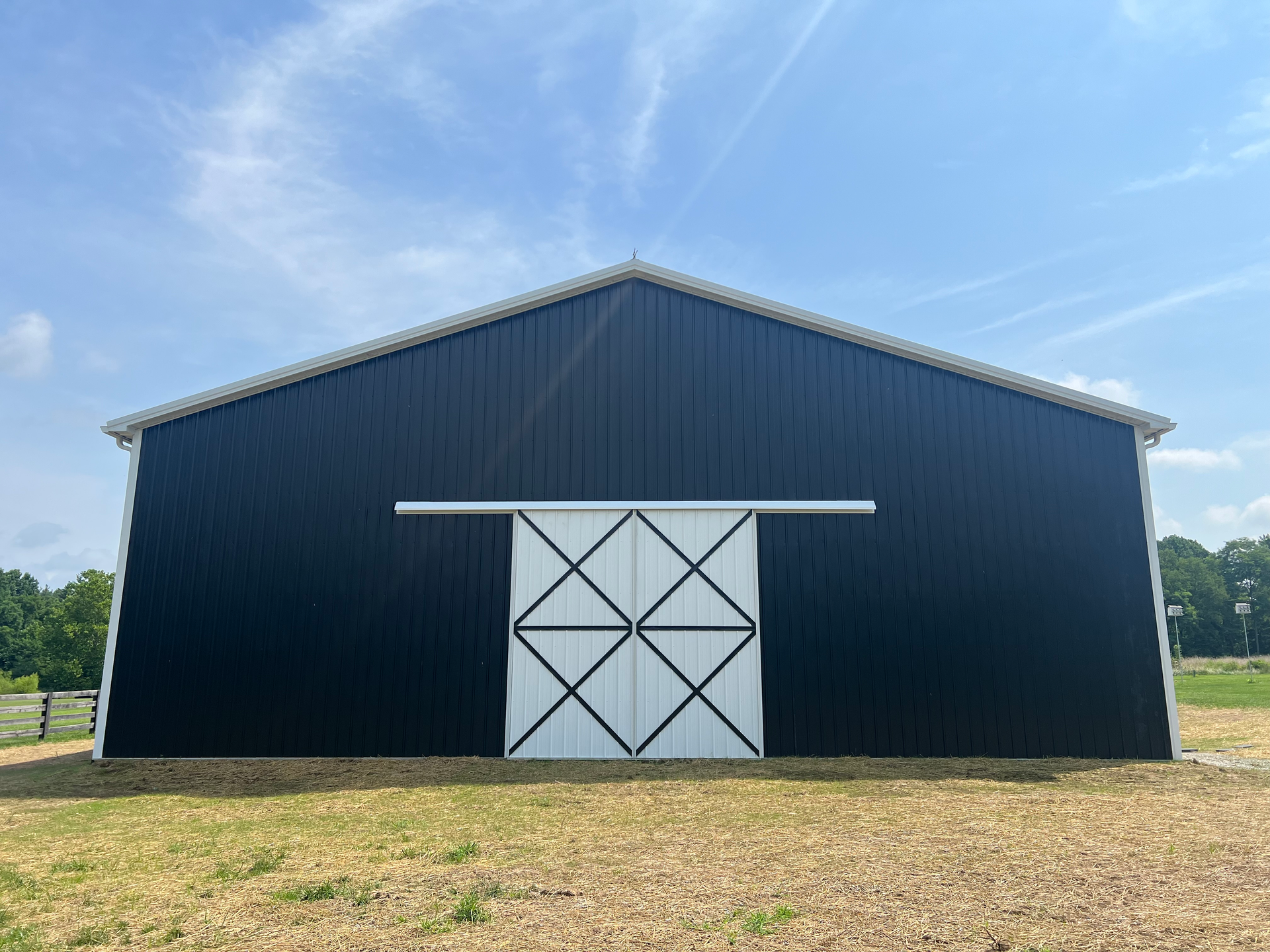 A black metal barn with a white sliding door featuring a black X-cross design stands in a grassy field under a blue sky.