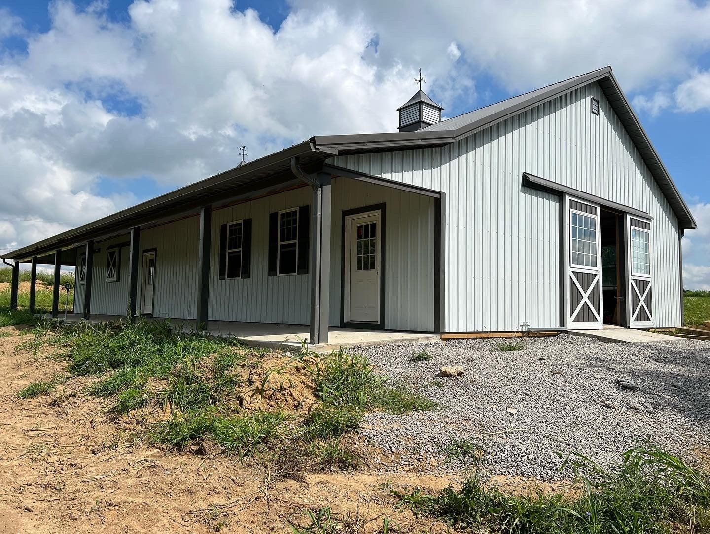 A light grey metal pole barn with a covered porch, a white entry door, and sliding barn doors under a bright, cloudy sky.