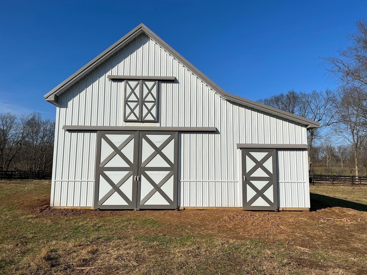 A white metal barn with dark gray trim and X-patterned doors sits in a grassy, open field under a clear blue sky.