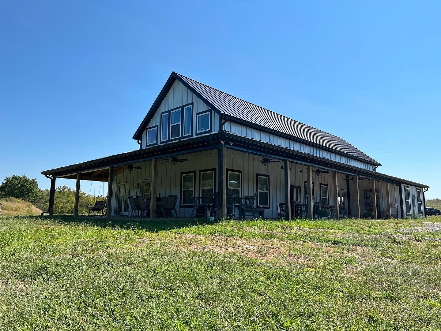 A white farmhouse with a large wraparound porch and dark trim sits on a grassy field under a clear blue sky.