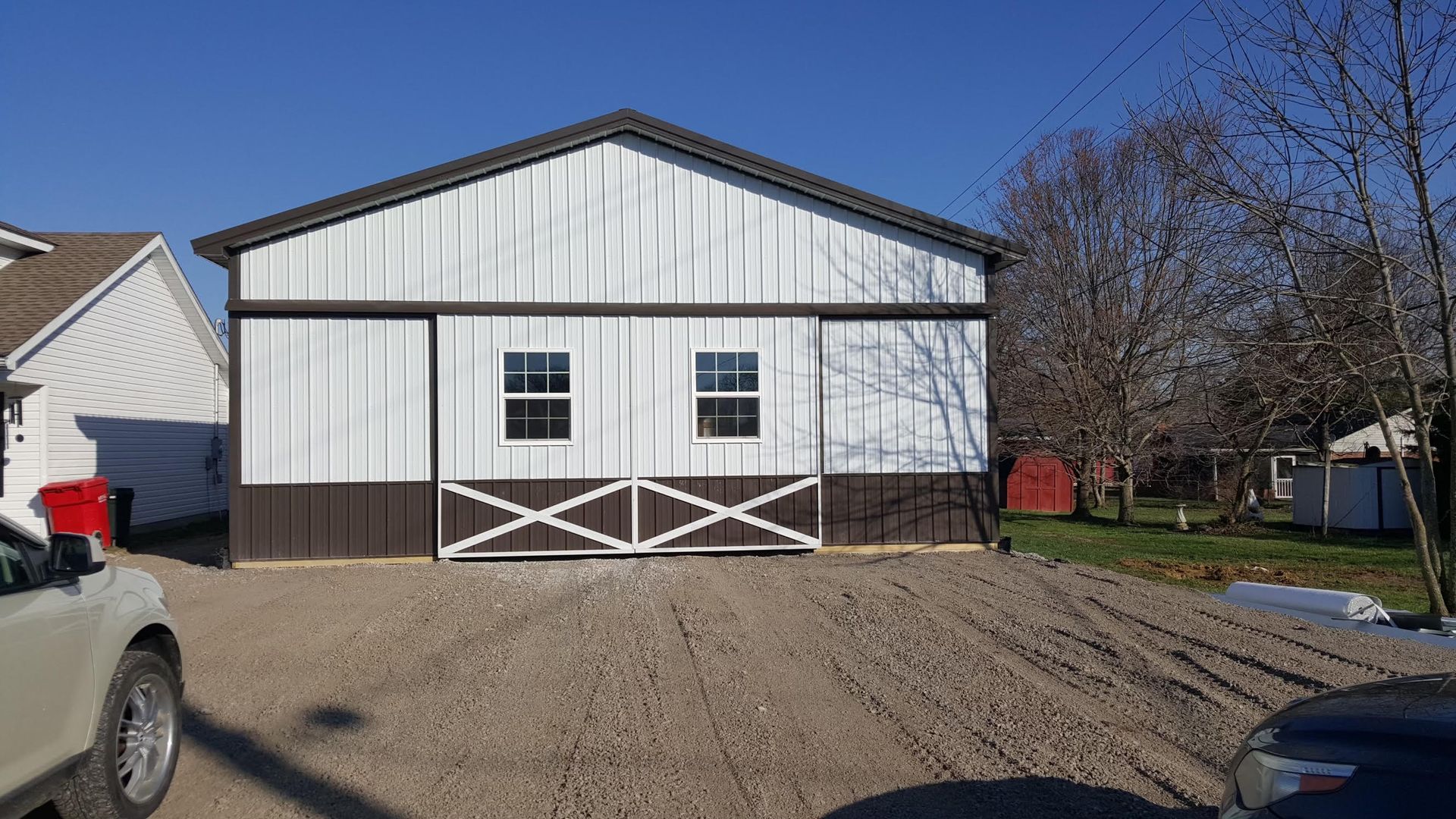 A white and brown metal garage building with a decorative X-pattern on the front, set on a gravel lot under a blue sky.