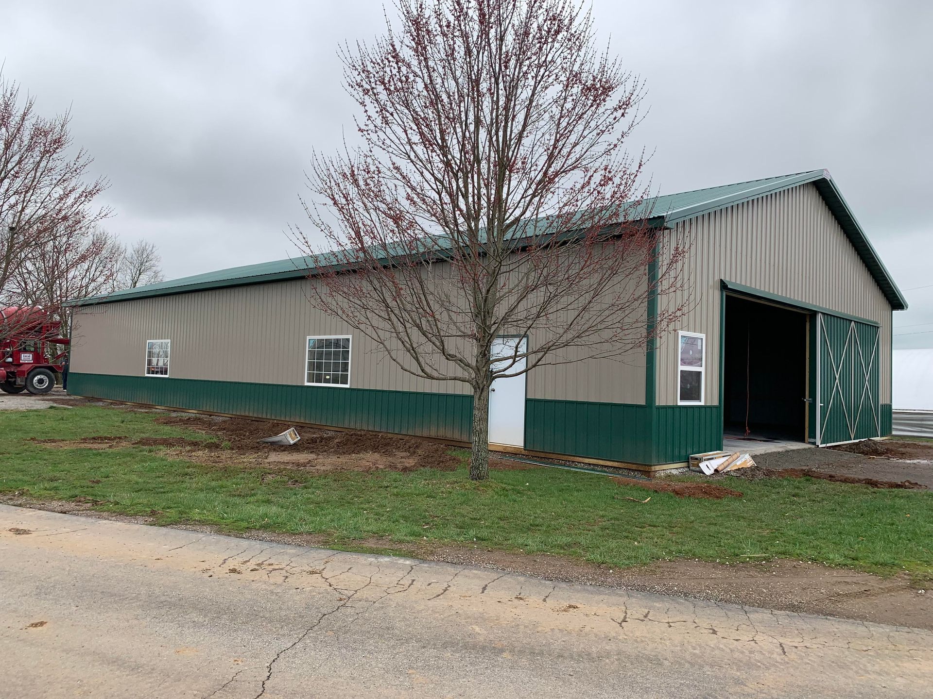 A tan and green metal barn with sliding doors and windows sits on a grassy lot next to a road under a cloudy sky.